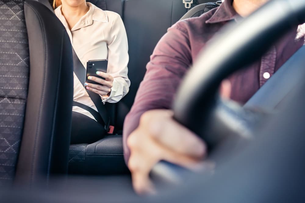 Una mujer de negocios se sienta en el asiento trasero de un taxi, con la atención fija en su teléfono móvil. El conductor se desplaza por las calles mientras la mujer se dedica a sus negocios sobre la marcha, confiando en una aplicación de viajes compartidos para desplazarse.