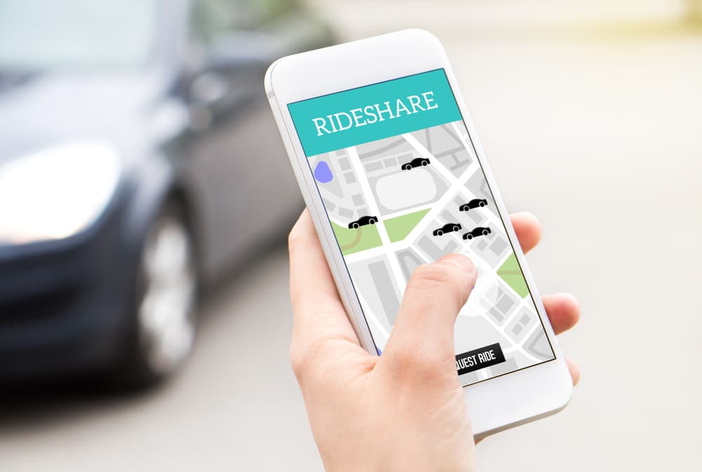 A woman holds her phone, its screen displaying a ride-sharing taxi service app. In the background, a car waits, symbolizing the convenience of ordering a ride with just a few taps on her mobile device.