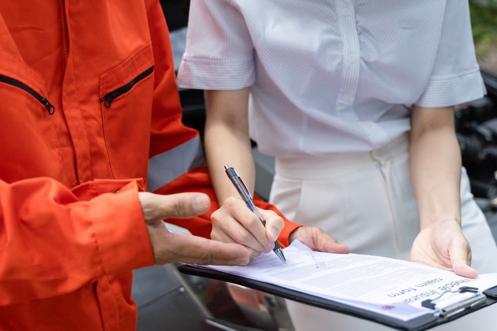 A person in a white shirt signing an insurance claim form held by someone in an orange jacket.