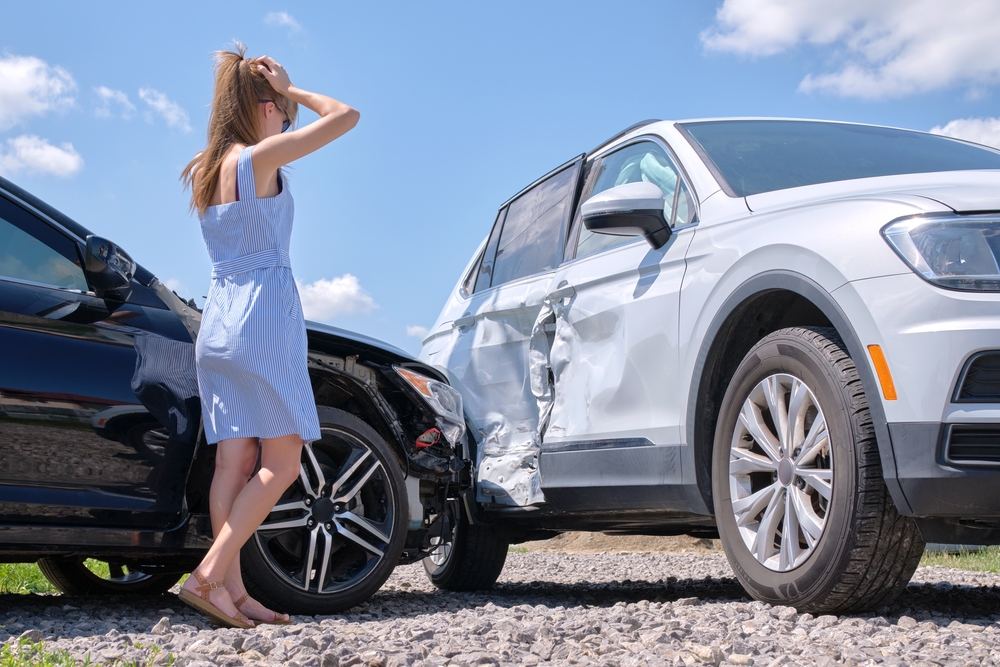 Sad young woman driver standing near her smashed car looking shocked after a left turn accident on a busy road.